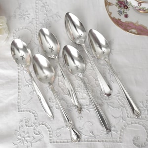 May include: A collection of silver spoons with various handle designs, arranged on a white embroidered tablecloth. A floral teacup and saucer are in the background, along with pink and white flowers. The spoons are reflective and elegant.
