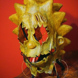 Könnte beinhalten: Eine handgefertigte, gelbe Maske mit einem gezackten, sonnenartigen Design. Die Maske hat genähte Nähte, einen klaffenden Mund und eine markante Nase. Schwarzer Draht und Lederriemen sind angebracht. Der Text "REDNECK SLASHER STUDIO" ist unten sichtbar.
