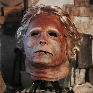 May include: A detailed, realistic, hand-painted mask of a man's face with a weathered, aged appearance. The mask is made of a brown material and has a white wig. The mask is mounted on a black tripod stand.