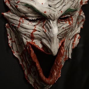 May include: A white and red mask with a grotesque, exaggerated smile. The mask is covered in blood splatters and has bandages on the nose and forehead. The mask is made of a textured material that looks like skin.