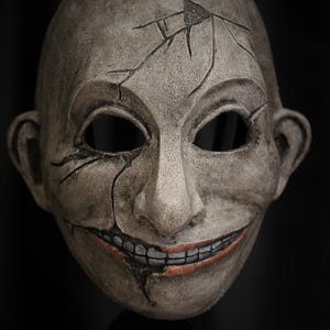 May include: A full-face mask with a weathered, cracked gray finish, featuring deep-set eye sockets, a prominent nose, and a wide, toothy grin with reddish-brown lips. The text REDNECK SLASHER STUDIO is printed in white in the bottom right corner.