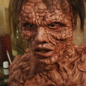 May include: Close-up of a detailed, realistic, red-toned, grotesque, and disturbing-looking monster mask. The mask has a stitched-up face with a wide open mouth and visible teeth.