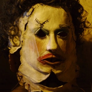 May include: A pale-faced mask with red lips and dark eye sockets, stitched together with visible seams. The mask has dark, curly hair and is mounted on a stand. The overall aesthetic is reminiscent of a horror movie prop.