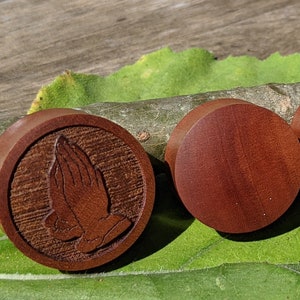 May include: Four wooden ear gauges with a praying hands design carved into the surface. Two of the gauges are plain wood, while the other two have the design.