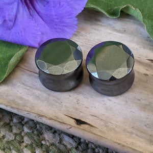 May include: Two black gemstone plugs with a faceted design. The plugs are on a wooden surface.