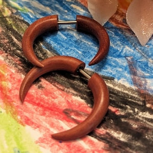 May include: Three reddish-brown wooden earrings with a curved, horn-like design. The earrings have a silver-colored post. The earrings are displayed on a colorful background with green and pink floral accents.