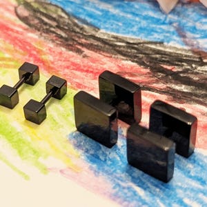 May include: A collection of black earrings in various geometric shapes. Two pairs of square stud earrings and two pairs of dumbbell-shaped earrings are displayed on a colorful background. The earrings have a glossy finish.