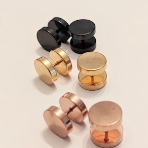 May include: An assortment of stud earrings in black, gold, and rose gold. The earrings have a flat, circular design and a post for insertion. The earrings are displayed on a white surface.