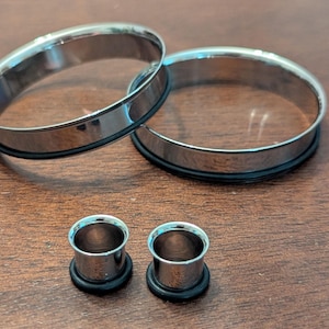 May include: Four silver-coloured ear gauges. Two larger gauges are circular with a black rubber ring. Two smaller gauges are cylindrical with a black rubber ring. All gauges are on a brown wooden surface.
