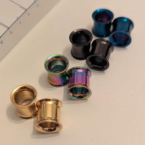 May include: A collection of body jewelry, including several ear gauges. The gauges are in various colors, including gold, black, and iridescent rainbow hues. The jewelry is cylindrical with a flared edge.