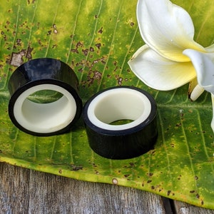 May include: Two black and white ear gauges sit on a green leaf with a white flower. The gauges are cylindrical with a black exterior and a white interior. The leaf has brown spots and is resting on a wooden surface.