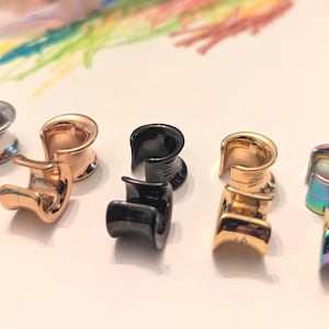 May include: An assortment of six metal ear gauges in various colors, including silver, rose gold, black, and iridescent finishes. The gauges are displayed on a light-colored surface, with a colorful abstract design in the background.