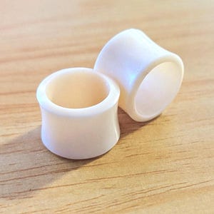 May include: A pair of off-white ear plugs with a smooth, concave saddle shape. These circular body jewelry pieces feature a solid, opaque finish and a hollow center, designed for stretched ear piercings with a flared edge on both sides.