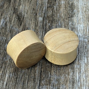 May include: Two wooden ear gauges, one is a flared style and the other is a straight style. Both are a light brown color.
