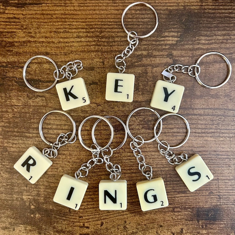 Puede incluir: Llaveros con fichas de Scrabble que deletrean la palabra "KEYS" y "RINGS". Cada ficha tiene una letra y un valor de puntos.