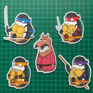 Puede incluir: Cinco pegatinas de dibujos animados con ping&uuml;inos vestidos como las Tortugas Ninja. Las pegatinas est&aacute;n sobre un fondo verde. Los ping&uuml;inos llevan bandanas rojas, azules, moradas y naranjas. Las pegatinas muestran las letras 'L', 'R', 'M' y 'D' en el pecho de los ping&uuml;inos.