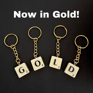 Puede incluir: Cuatro llaveros dorados con fichas de Scrabble que deletrean la palabra "GOLD". Cada ficha tiene un valor de puntos diferente.