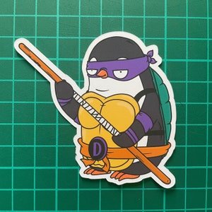 Puede incluir: Un ping&uuml;ino de dibujos animados con un disfraz de tortuga ninja verde y morado, sosteniendo un bast&oacute;n de madera. El ping&uuml;ino lleva una concha amarilla con una "D" negra.