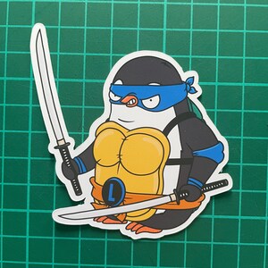 Puede incluir: Un ping&uuml;ino de dibujos animados con un disfraz de tortuga ninja azul y negro con un caparaz&oacute;n amarillo y una espada blanca. El ping&uuml;ino sostiene una espada blanca en cada mano. El ping&uuml;ino lleva una bandana azul con la letra "L" en ella.