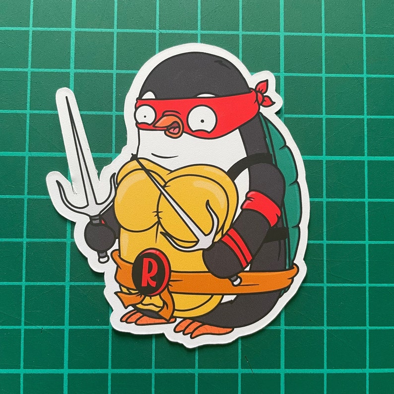 Puede incluir: Un ping&uuml;ino de dibujos animados con un pa&ntilde;uelo rojo y un caparaz&oacute;n de tortuga amarillo y naranja con una 'R' roja en la parte delantera. El ping&uuml;ino sostiene un sai en cada mano.