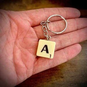 Puede incluir: Un llavero de baldosa de Scrabble blanco con la letra "A" y el n&uacute;mero "1". La baldosa est&aacute; unida a un llavero plateado.