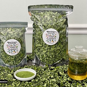 May include: Two bags of dried moringa leaves, one large and one small, with a label that reads "Moringa Leaves Yard Yummmies". A small white bowl with moringa powder and a glass mug of moringa tea are also shown.