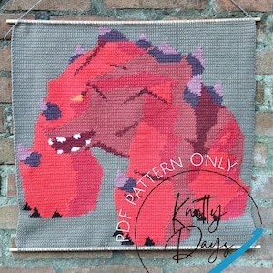 Può includere: Arazzo da parete all'uncinetto con un disegno di dinosauro rosso e rosa su sfondo grigio. Il testo "PDF PATTERN ONLY" e "Knotty Days" sono visibili. Appeso a un'asta di legno.