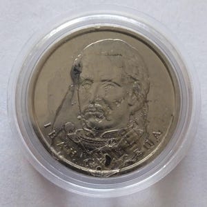 Puede incluir: Una moneda de plata dentro de un soporte de plástico transparente. La moneda presenta un retrato detallado de un hombre con bigote y chaqueta de estilo militar. La superficie de la moneda está desgastada.