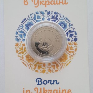 Puede incluir: Una moneda de plata con un diseño de un bebé en posición fetal. La moneda está en una caja de plástico transparente. El texto en la moneda dice "Народжений в Україні", que se traduce como "Nacido en Ucrania". El texto en la tarjeta dice "Nacido en Ucrania" y "Banco Nacional de Ucrania".