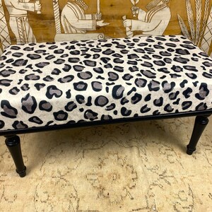 May include: A rectangular ottoman with a black frame and legs. The cushioned top is covered in a cream-colored fabric with a pattern of black and gray leopard spots. The ottoman is a decorative furniture piece.