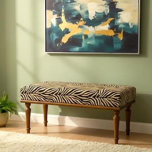 Puede incluir: Banco rectangular con cojín con estampado de cebra y patas de madera. El banco está colocado contra una pared verde claro, con una pintura encima. Una pequeña planta en maceta está a la izquierda.