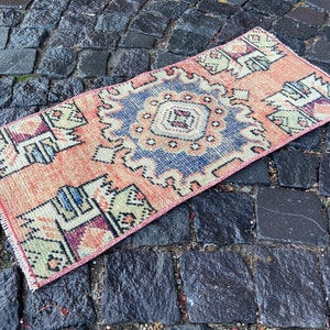 May include: A rectangular, hand-woven rug with a central medallion design. The rug features a peach-coloured field with a blue and cream-coloured central design. Geometric patterns in green, purple, and blue adorn the borders. The rug is placed on a cobblestone surface.
