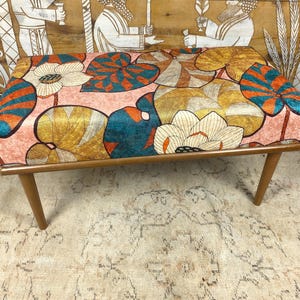 May include: A vintage-style bench with a floral design in pink, orange, teal, and yellow. The bench has a rectangular seat and four wooden legs. The floral pattern features large stylized flowers and leaves. The bench is approximately 90 cm long.