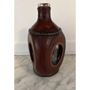May include: A brown leather-covered bottle with a clear glass stopper. The bottle features two oval cutouts showcasing the glass interior. Decorative stitching enhances the design, adding a touch of elegance to this unique item.