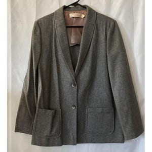 May include: A grey blazer with two front pockets and two buttons. The blazer has a notched lapel and is hanging on a black hanger. The interior lining is a light brown colour. The blazer is made of a wool-like material.