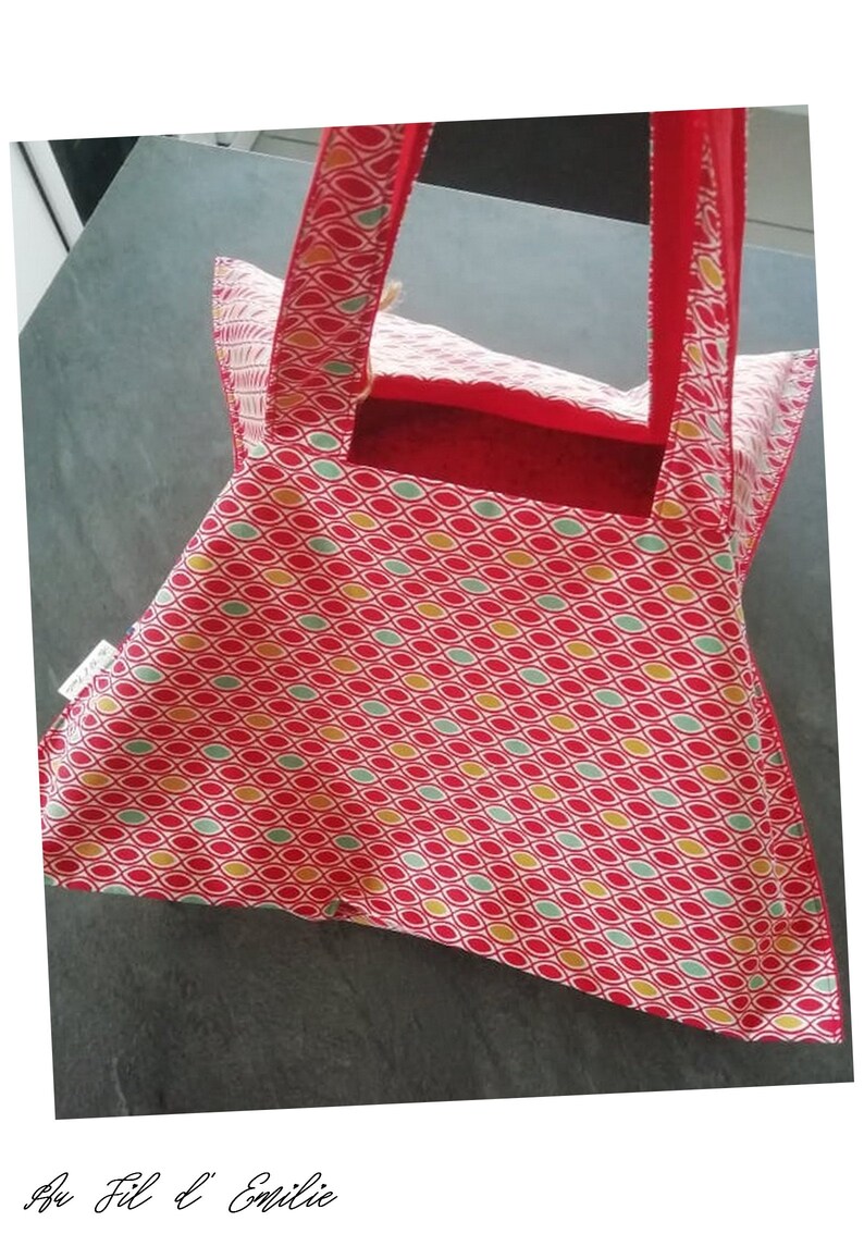 May include: Red and white tote bag with a geometric pattern of green, yellow, and red ovals. The bag has red handles and a red lining. The bag is made of fabric and has a label that says "Au Fil d'Emilie".
