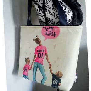 Op de afbeelding: Een witte tote bag met een kleurrijke illustratie van een vrouw en twee kinderen. De vrouw draagt een roze shirt met "QUEEN 01" op de rug. De kinderen dragen roze en zwarte shirts met "01" op de rug. De tas heeft een roze spraakballon met de tekst "Mom life is the best life".