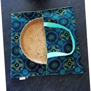 May include: A teal and yellow patterned fabric tote bag with a brown ceramic plate inside. The bag has two teal handles.