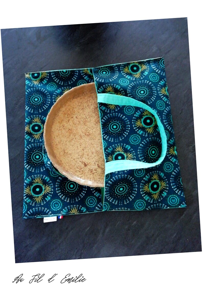 May include: A teal and yellow patterned fabric tote bag with a brown ceramic plate inside. The bag has two teal handles.