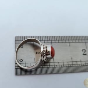 Może przedstawiać: Srebrny pierścionek z czerwonym kamieniem szlachetnym osadzonym w oprawie typu bezel. Rozmiar pierścionka jest oznaczony na linijce i wynosi 32.