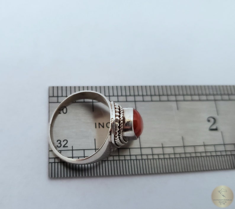 Może przedstawiać: Srebrny pierścionek z czerwonym kamieniem szlachetnym osadzonym w oprawie typu bezel. Rozmiar pierścionka jest oznaczony na linijce i wynosi 32.