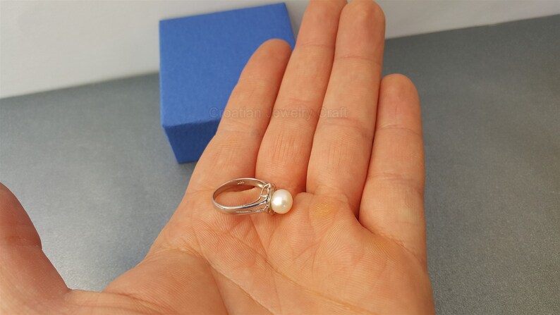 May include: A silver ring with a white pearl set in a silver band. The ring is on a hand.