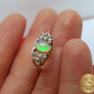 May include: A silver ring with a large oval opal gemstone surrounded by smaller clear gemstones. The opal has a rainbow of colors, including green, pink, and purple.