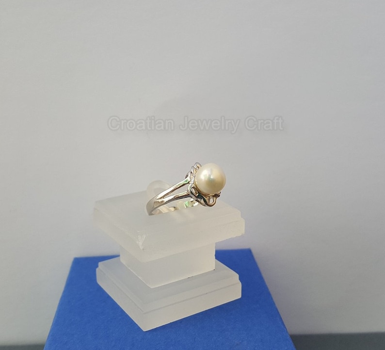 May include: A silver ring with a white pearl set in a delicate, intricate design. The ring is displayed on a white pedestal.
