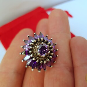 May include: A silver ring with a purple gemstone surrounded by smaller purple gemstones and a silver filigree design.