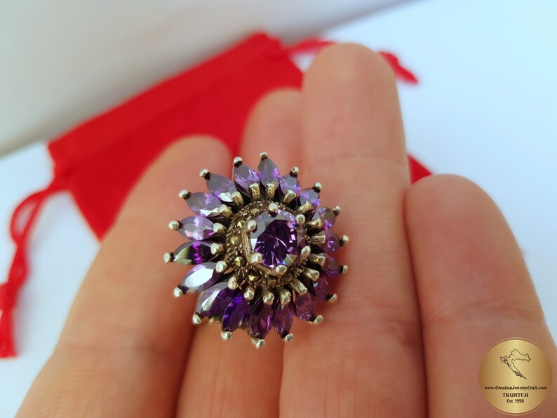 May include: A silver ring with a purple gemstone surrounded by smaller purple gemstones and a silver filigree design.