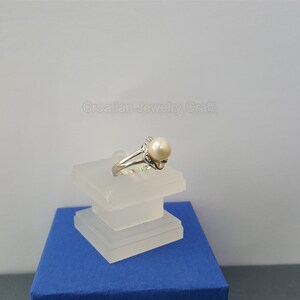 May include: A silver ring with a white pearl set in a delicate floral design. The ring is displayed on a white stand.