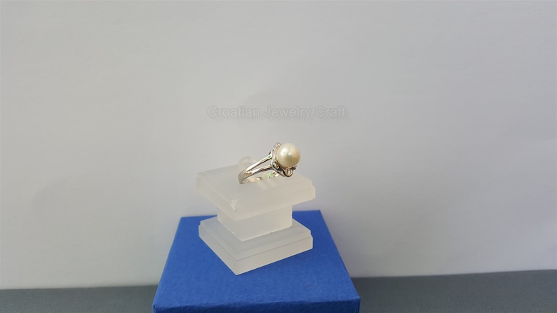 May include: A silver ring with a white pearl set in a delicate floral design. The ring is displayed on a white stand.