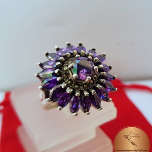 May include: A silver ring with a large purple gemstone surrounded by smaller purple gemstones. The ring is set in a flower-like design.