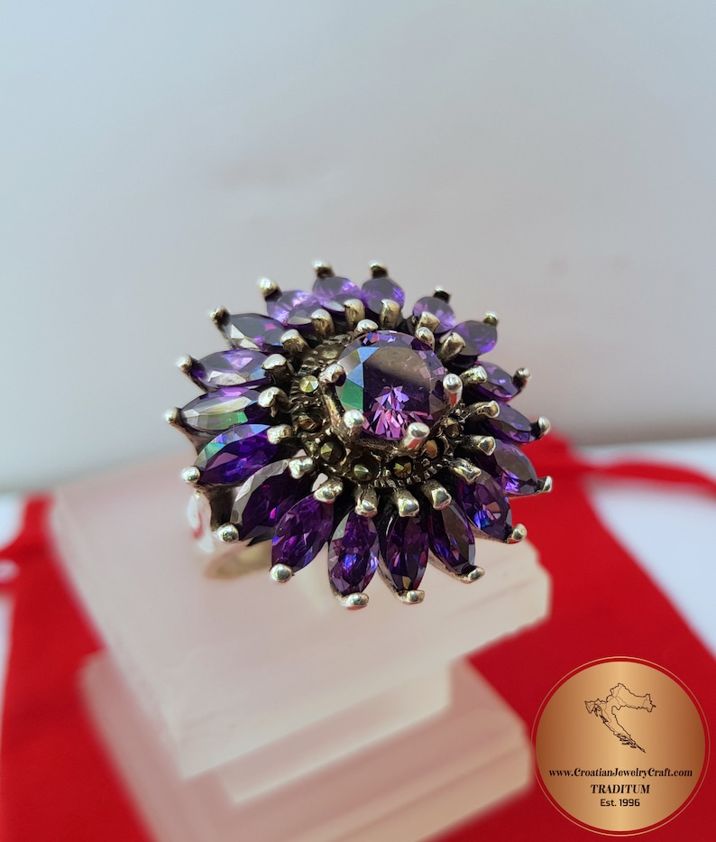 May include: A silver ring with a large purple gemstone surrounded by smaller purple gemstones. The ring is set in a flower-like design.